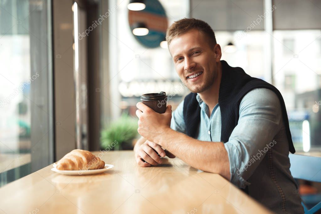 young handsome man eating his croissant and drinking hot coffee for lunch in the cafe feeling happy