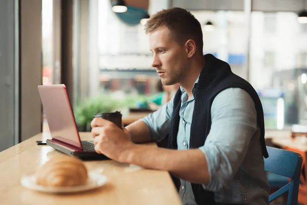 Joven hombre guapo trabaja en su computadora portátil en la cafetería comiendo croissant y beber café caliente para el almuerzo — Foto de Stock