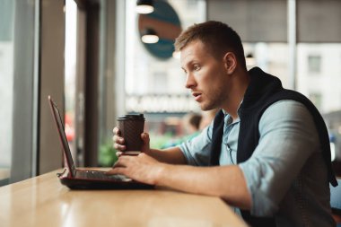 Uzaktan kumandalı bilgisayarında çalışan yakışıklı genç adam kafedeki sıcak kahveyi görünce şaşırmış görünüyor.