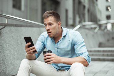 Akıllı telefon tutan ve öğle tatilinde kahve içen genç yakışıklı ofis çalışanı sokakta merdivenlerde otururken şaşırmış