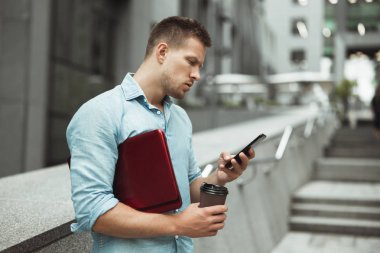 genç yakışıklı ofis çalışanı öğle tatili sırasında sokakta duran dizüstü bilgisayar ve kahve fincan tutan onun smarphone kontrol