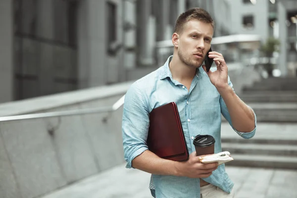 young handsome office worker talking on phone holding laptop cup of coffee and sandwich walking down the street near business center eating on the go