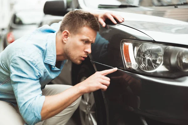 Joven hombre guapo sentado cerca de su coche encontró algunos arañazos en su parachoques buscando sorprendido — Foto de Stock