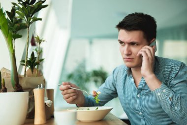 genç yakışıklı adam ofis yakınındaki kafede mola sırasında salata ve öğle yemeği için kahve içerken telefonda konuşuyor