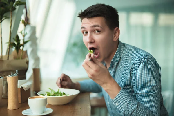 Joven guapo hombre con hambre come ensalada y bebe café para el almuerzo durante el descanso en la cafetería cerca de la oficina — Foto de Stock