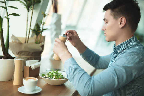 Jovem bonito homem salgando salada preparando-se para comer o seu almoço e bebe café durante o intervalo no café perto do escritório — Fotografia de Stock