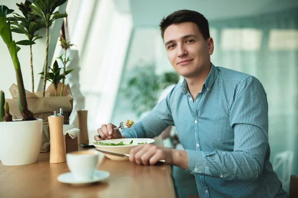 Joven guapo feliz hombre come ensalada y bebe café para el almuerzo durante el descanso en la cafetería cerca de la oficina — Foto de Stock