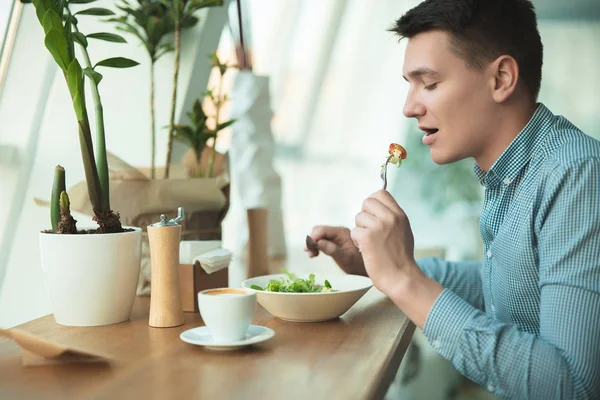 Jonge knappe man eet salade en drinkt koffie voor de lunch tijdens pauze in café in de buurt van Office ziet er hongerig uit — Stockfoto