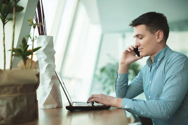 Joven hombre de negocios guapo tiene conversación en su teléfono inteligente y trabaja en su computadora portátil mientras espera el almuerzo en la cafetería cerca de la oficina — Foto de Stock