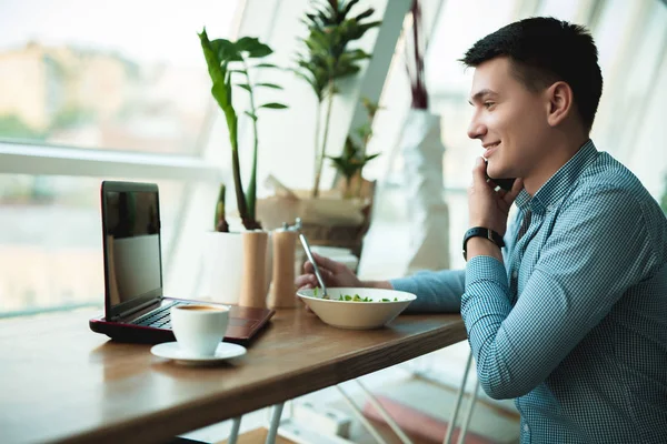 young handsome smiling businessman drinks his hot coffee and eats salad for lunch while working in his laptop during break at trendy cafe near office