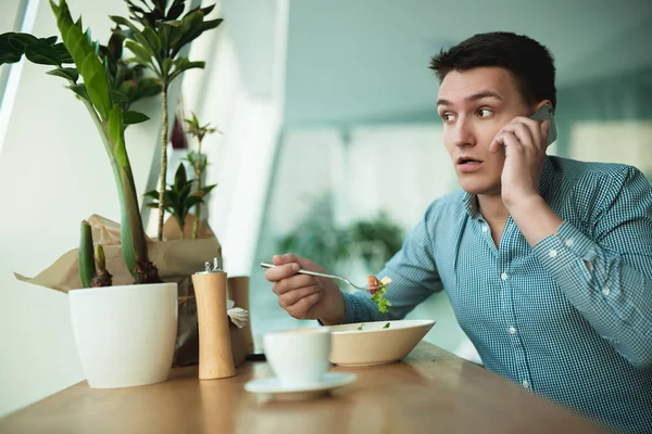 young handsome man speaks on the phone while eating salad and drinking coffee for lunch during break at cafe near office looks worried