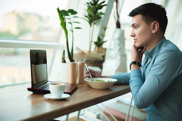 young handsome businessman drinks his hot coffee and eats salad for lunch while working in his laptop during break at trendy cafe near office