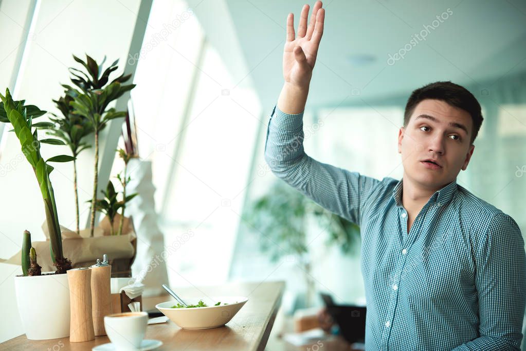young handsome man calls waiter raising his hand while eating salad and drinking coffee for lunch during break at cafe near office