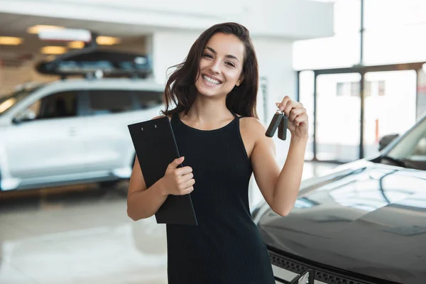 Bella bruna donna manager sorridente tenuta brochure in una mano e chiavi dell'auto in un altro in piedi nel centro di distribuzione — Foto Stock