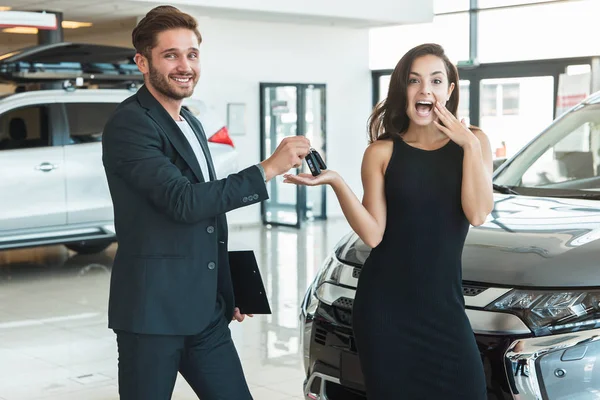 handsome man manager giving car keys to beautiful brunette woman client standing near brand new car after succesful deal in dealership center feeling happy