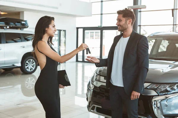 Hermosa morena gerente de la mujer dando llaves del coche al hombre guapo cliente de pie cerca de nuevo coche después de un acuerdo exitoso en el centro de concesionarios sentirse feliz — Foto de Stock