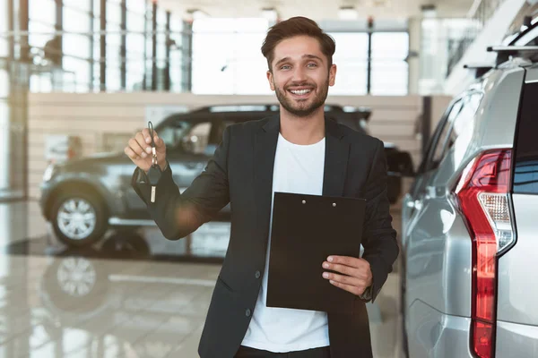Молодой красавец-менеджер улыбается с брошюрой в руке, стоя в дилерском центре рядом с новым внедорожником с ключами от машины — стоковое фото