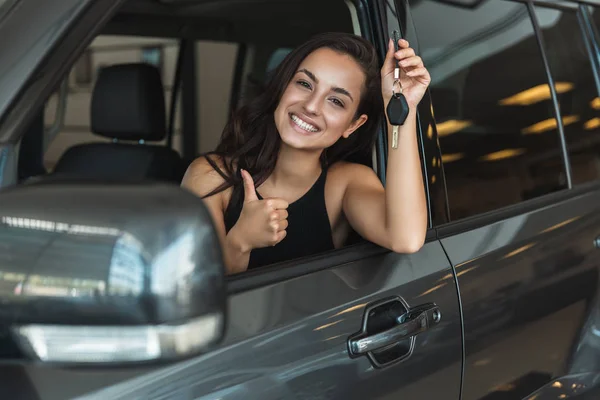 Beautiful brunette woman sitting in brand new car after succesful deal in dealership center feeling happy holding car keys showing like sign — Stock Photo, Image