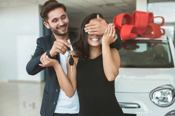 Jeune bel homme fait surprise pour sa belle femme ferme les yeux avec ses mains debout dans le centre de concession acheter une nouvelle voiture pour le présent — Photo