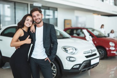 güzel genç mutlu çift erkek ve kadın tabela gibi gösteren bayilik merkezinde duran yepyeni bir araba seçerek