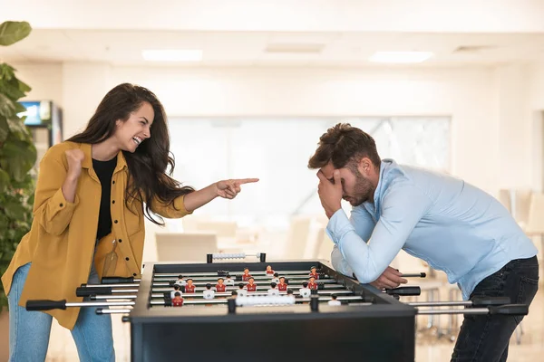 Dua rekan kerja berambut cokelat wanita cantik dan pria muda tampan bermain sepak bola meja selama istirahat di kantor, dia menang dan tertawa dan dia marah — Stok Foto