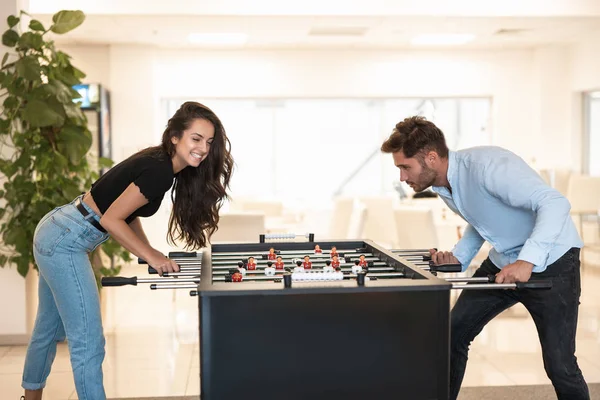 Dua rekan muda berambut cokelat wanita cantik dan pria tampan bermain sepak bola meja selama istirahat di kantor tampak terkonsentrasi pada permainan — Stok Foto