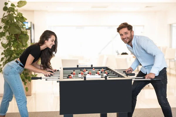 Dos jóvenes colegas morena hermosa mujer y hombre guapo jugando futbolín durante el descanso en la oficina buscando involucrado en el juego —  Fotos de Stock