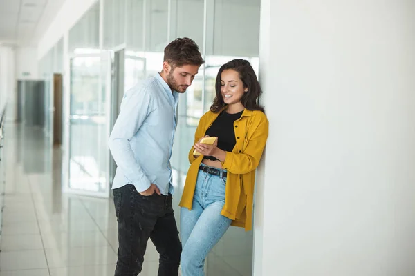 Två unga kollegor stilig man och vacker kvinna flirta tillsammans på kontoret hon visar honom något i smartphone — Stockfoto