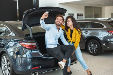 güzel genç çift erkek ve kadın açık araba bagajında oturan selfie çekim bayilik merkezinde yeni bir araba arıyor