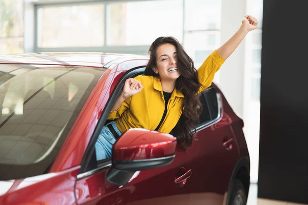 Bella donna bruna in cerca di nuova auto nel centro di concessionaria che sporge fuori dal finestrino del veicolo felicemente — Foto Stock