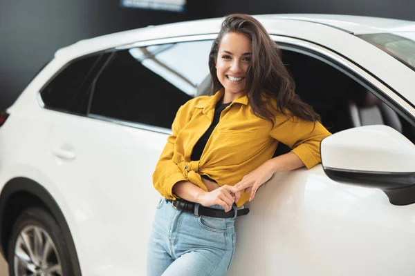 Bella donna bruna in cerca di nuova auto nel centro di concessionaria appoggiato sul veicolo bianco cercando felice — Foto Stock