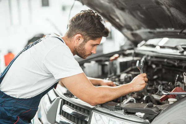 Joven mecánico guapo sonriente en uniforme la fijación de los problemas del motor en el capó del coche que trabaja en el centro de servicio — Foto de Stock