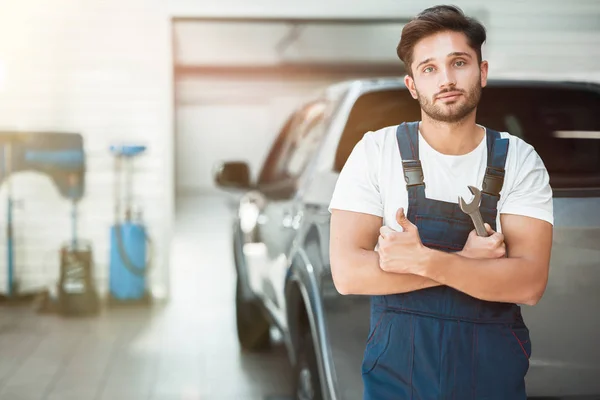 Joven mecánico guapo vistiendo uniforme celebración de la llave de pie en el centro de servicio de coches que muestra como signo — Foto de Stock