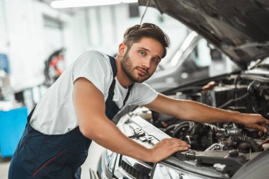 Genç gülümseyen yakışıklı tamirci üniforma giyiyor araba kaputunda motor tamir ediyor servis merkezinde çalışıyor mutlu görünüyor