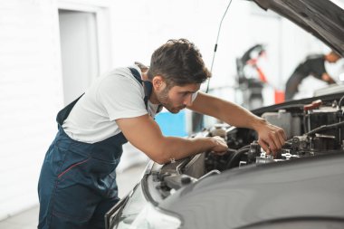 Üniformalı, gülümseyen genç tamirci araba kaputunda motor tamir ediyor servis bölümünde çalışıyor.