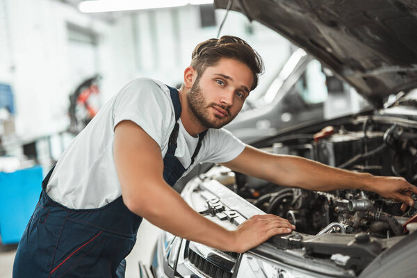 young smiling handsome mechanic wearing uniform fixing motor in car bonnet working in service center looking happy