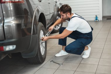 Araba servisinde çalışan, üniforma giyen genç yakışıklı tamirci İngiliz anahtarıyla lastiği tamir ediyor.