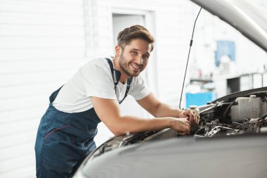 Genç, yakışıklı, gülümseyen, üniforma giyen araba kaputunda motor tamir eden tamirci servis bölümünde çalışıyor.