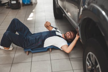 Araba servisinde çalışan üniformalı genç yakışıklı tamirci İngiliz anahtarı yardımıyla araç şasesini tamir ediyor.
