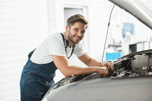 Jeune beau mécanicien souriant portant un moteur de fixation uniforme dans le capot de voiture travaillant dans le département de service — Photo