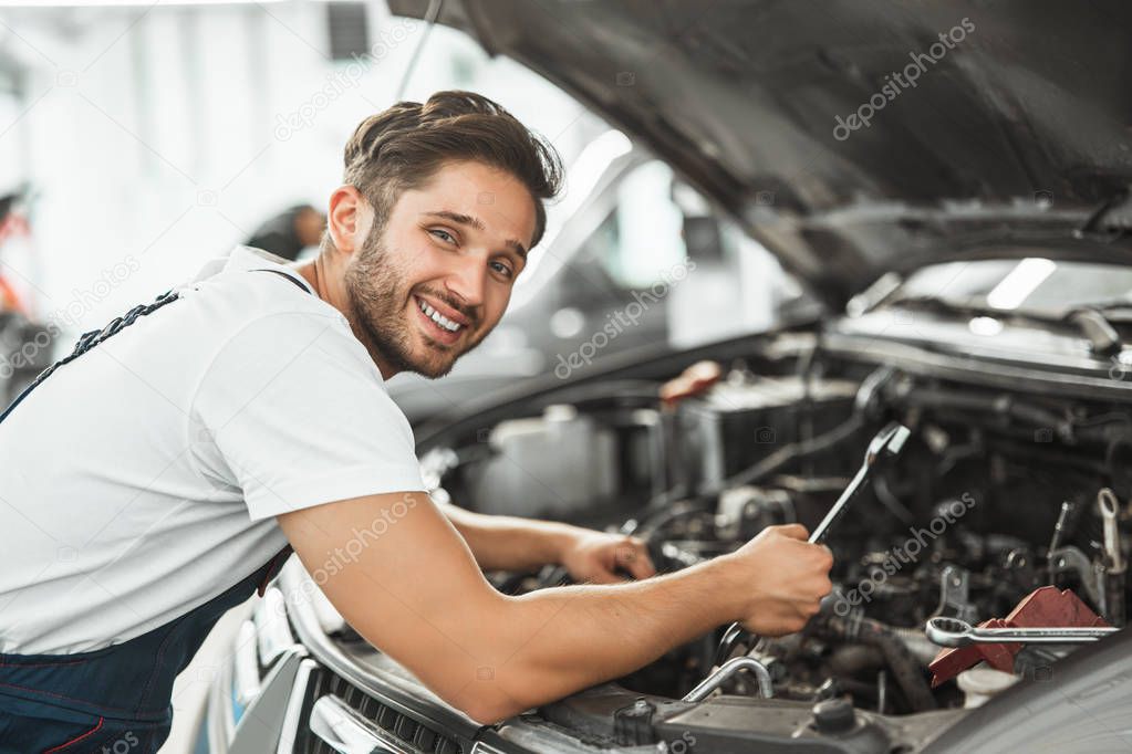 young smiling mechanic in uniform fixing motor problems in car bonnet working in service center