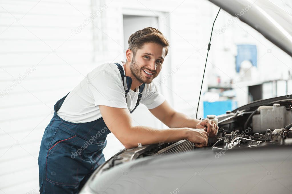 young handsome smiling mechanic wearing uniform fixing motor in car bonnet working in service department