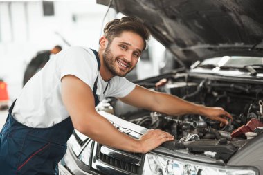 Üniformalı yakışıklı genç tamirci servis merkezinde çalışan araba kaputunda motor tamir ediyor.