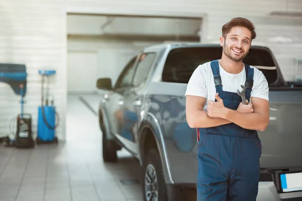 Joven mecánico sonriente guapo en uniforme la celebración de la llave de pie en el centro de servicio de coches que muestra como signo — Foto de Stock