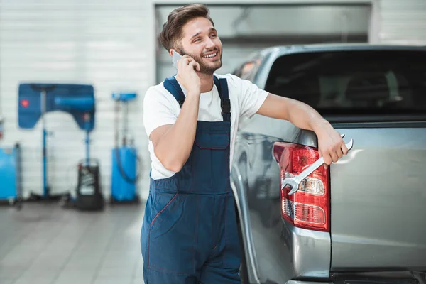 Joven mecánico guapo con uniforme apoyado en el maletero de recogida hablando por teléfono en el centro de servicio de coches — Foto de Stock