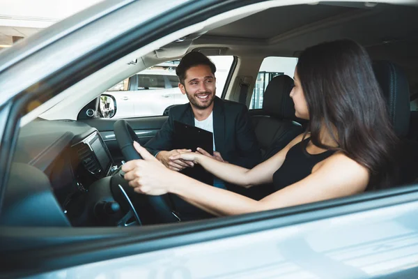 Przystojny mężczyzna i młoda piękna kobieta sprawdzanie nowe wnętrze samochodu siedząc w nowym samochodzie w centrum dealerskim — Zdjęcie stockowe