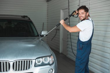 Üniformalı gülümseyen genç adam araba yıkama istasyonunda yüksek basınçlı su kullanarak araba yıkıyor.