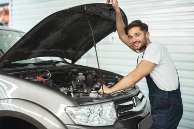 Üniformalı genç gülen tamirci servis merkezinde çalışan açık araba kaputunun yanında duruyor.