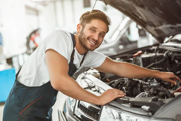 Joven mecánico guapo sonriente en el motor de fijación uniforme en el capó del coche que trabaja en el centro de servicio mirando feliz — Foto de Stock