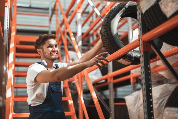 Joven mecánico guapo en uniforme que trabaja en el departamento de servicio de coches poniendo neumáticos para el almacenamiento — Foto de Stock